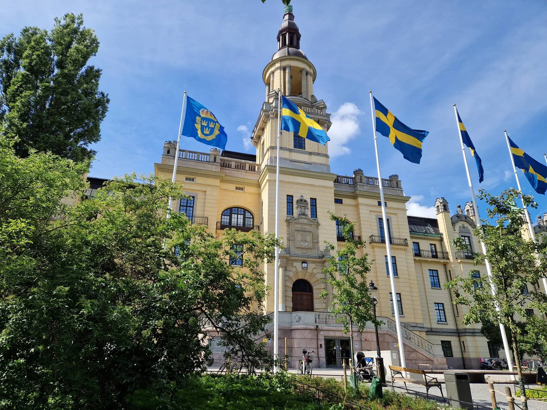 Gul byggnad med centralt torn, flera svenska flaggor, träd och parkbänk under blå himmel