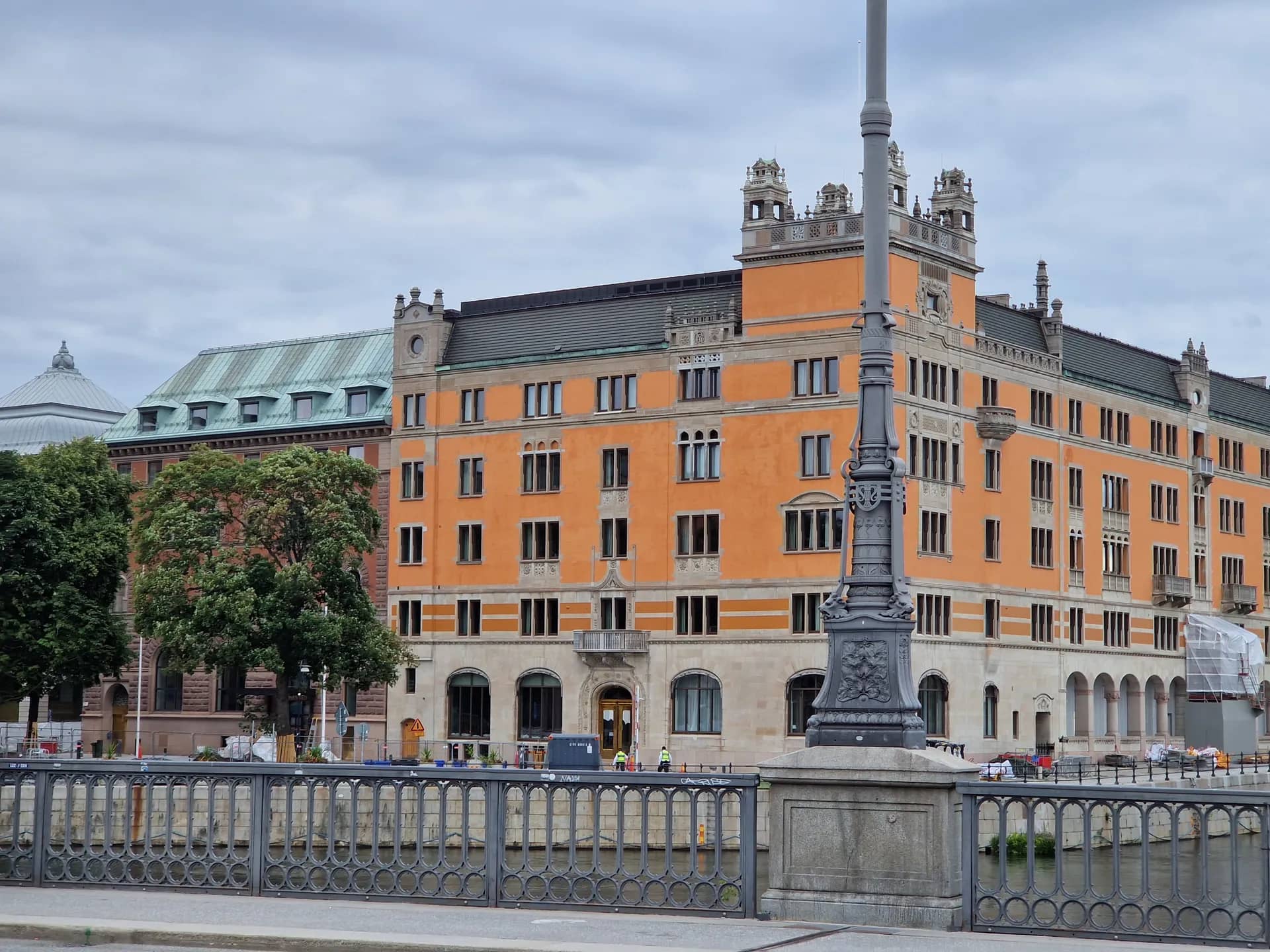 Stort orange hus med mörkt tak, prydda torn och många fönster. I förgrunden ett grått räcke vid vatten och en stor grå pelare