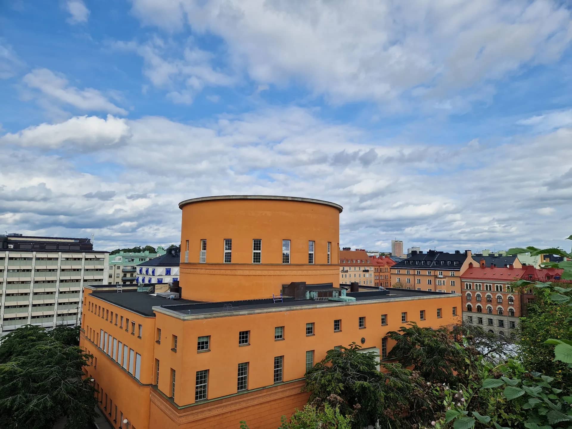 Orange byggnad med rund topp och fönster. Omkringliggande hus och grönska under en molnig himmel