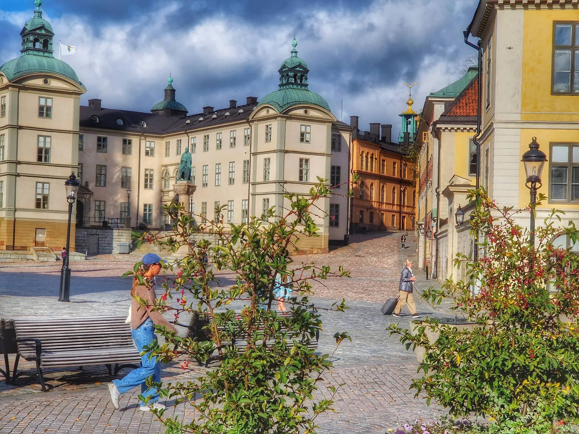 Historiskt torg med ståtliga byggnader med gröna kupoler, staty och gatsten. Molnig himmel. Folk går runt buskar med röda bär