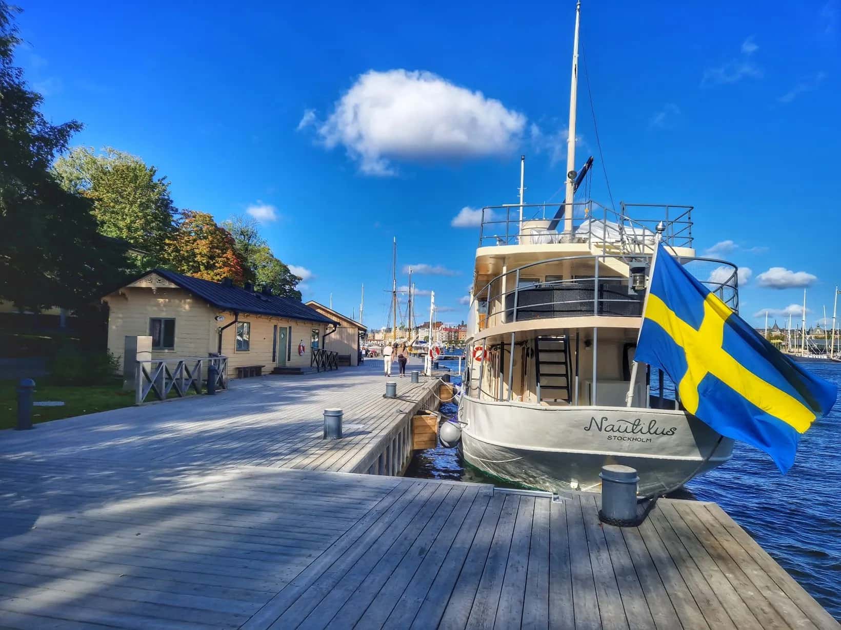 Trädäck vid vattnet med båten Nautilus Stockholm och svensk flagga. Gula hus, träd och blå himmel med moln