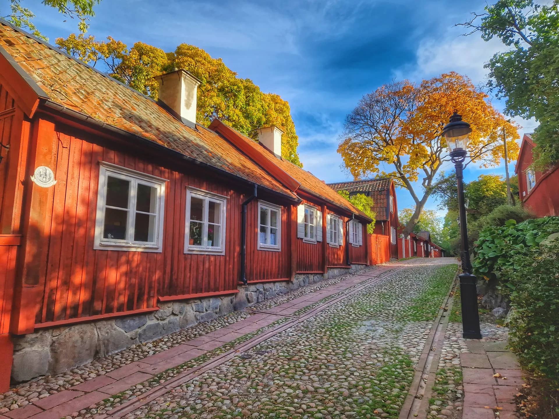 Röda trähus med vita fönster längs en kullerstensgata under höstträd och blå himmel