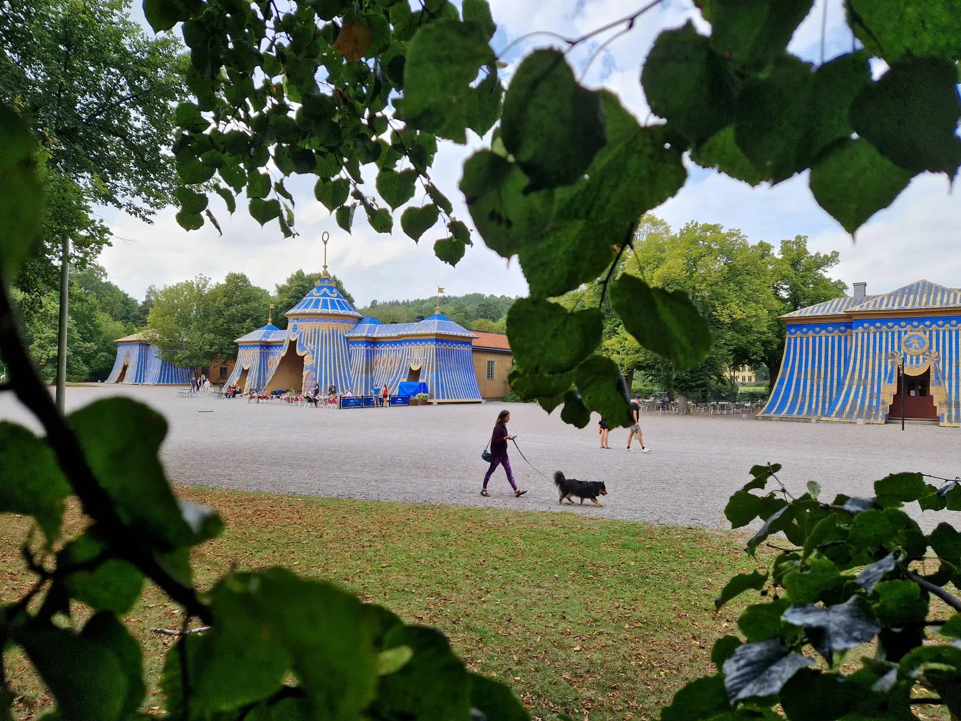 Blågulrandiga paviljonger på grusplan, med träd, himmel och en kvinna som går med hund. Löv i förgrunden