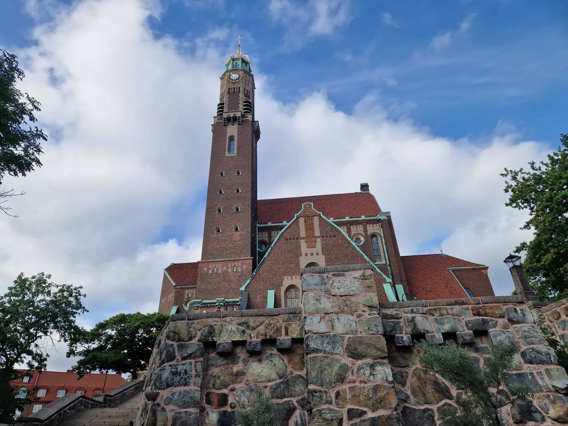 Brun tegelkyrka med ett högt torn, rött tak och gröna koppardetaljer. En stenmur i förgrunden, blå himmel med moln