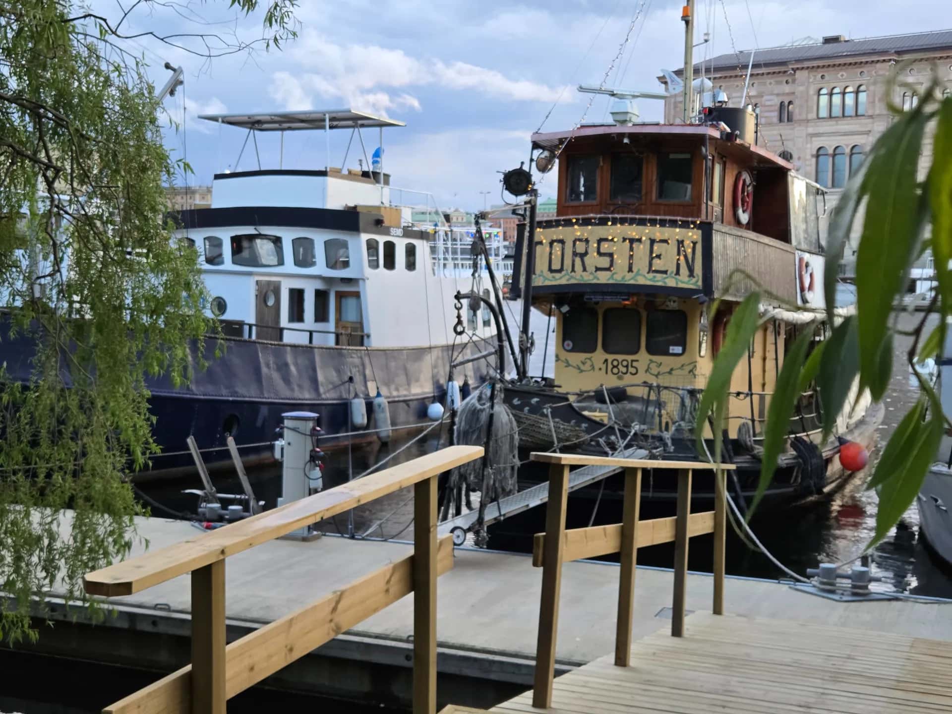 Två båtar vid en träbrygga med räcke. Den ena båten har namnet TORSTEN och årtalet 1895. Gröna löv inramar bilden