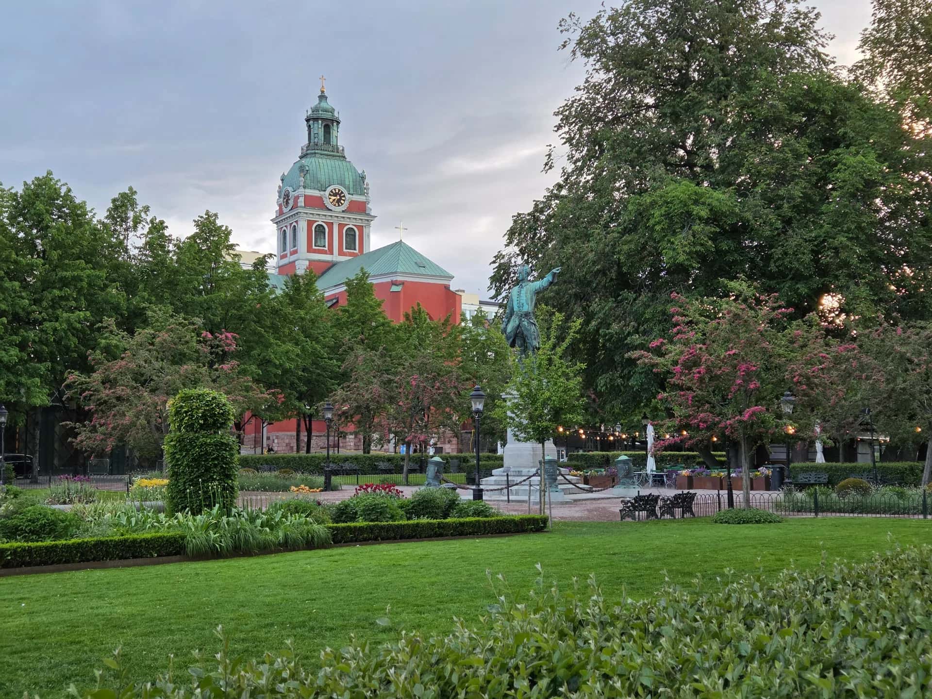 Park med grön gräsmatta, träd och blommande buskar. I bakgrunden syns en röd kyrka med grön kupol och ett klocktorn samt en staty