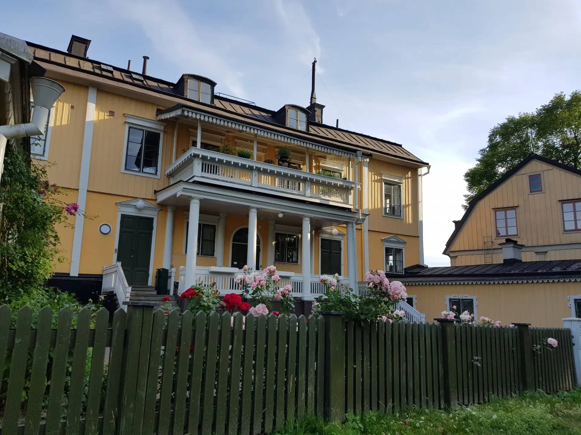 Ljusgult trähus med vit balkong, mörkt tak och ett grönmålat staket med rosor. Blå himmel med moln