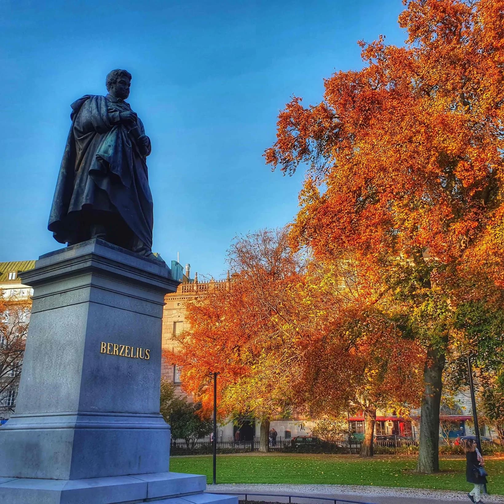 Mörk staty av en man på stenpelare med texten BERZELIUS, mot blå himmel och sprakande höstlövsträd